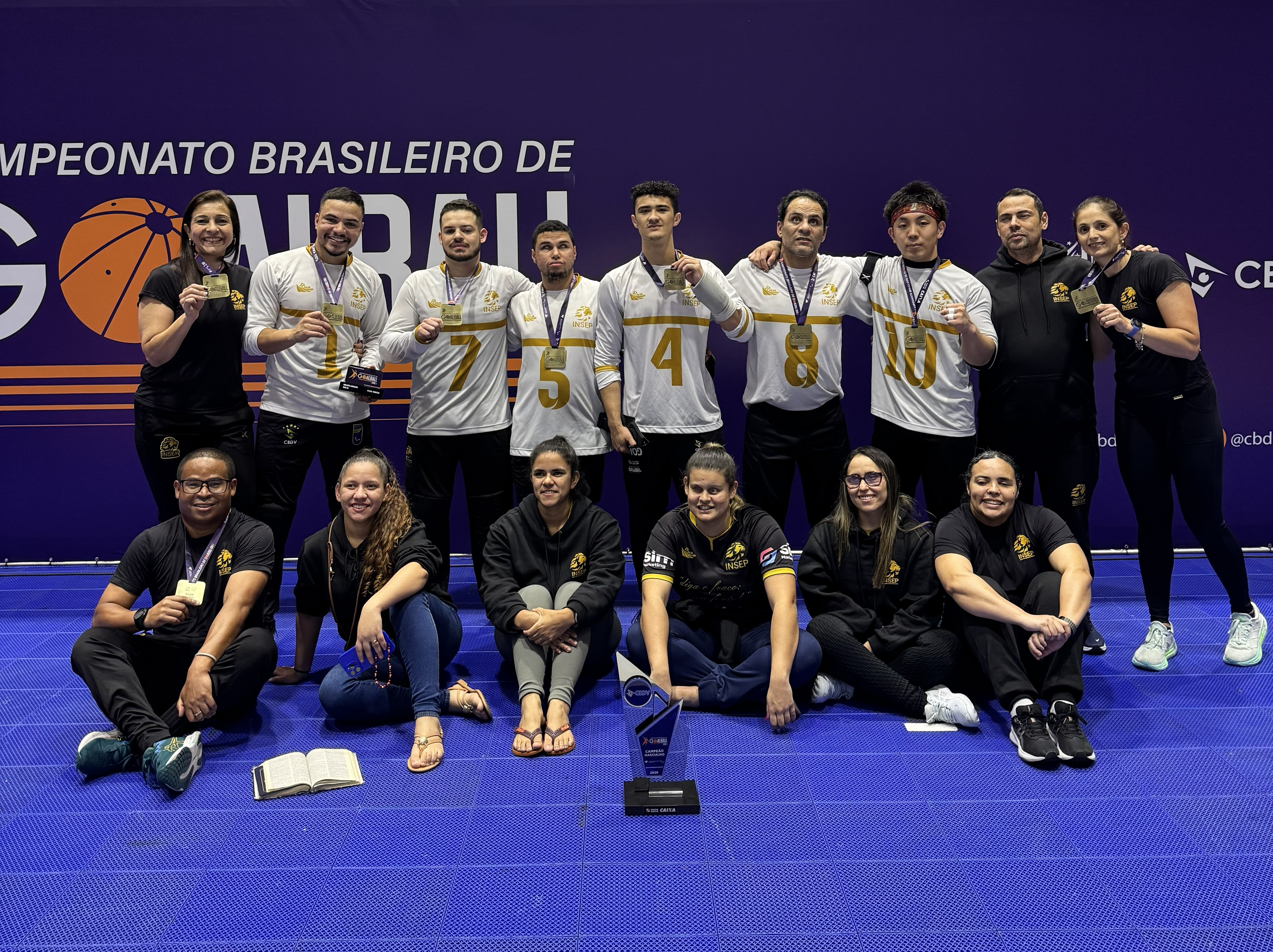 Atletas da equipe de Goalball do INSEP comemoram vitória em quadra, com medalhas no pescoço e troféu na mão.